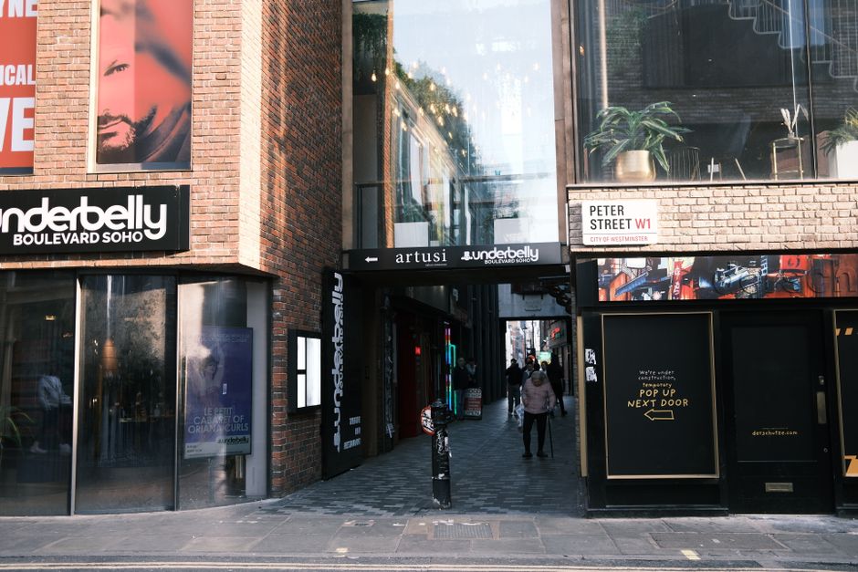 The image shows the entrance to an urban shopping and entertainment area on Soho's Carnaby Street, with the exterior of the Undertbelly venue visible on the left, featuring large glass windows and a brick facade with signage. To the right, there is a narrow pedestrian walkway leading into a passageway framed by a black awning with the name 'artusi,' and several people walking through the street, some carrying shopping bags or backpacks. The area is bustling with activity, and the environment includes modern buildings with large windows, indoor plants visible inside the upper floors, and signs indicating nearby businesses. The scene captures the process of city-based home relocation or furniture transport, with visible movement and the setting for a typical moving or packing scenario outside a commercial property in Soho, where professional movers like Man with Van Soho may operate. Equipment such as bollards and pavement details are also visible, emphasizing the urban context.