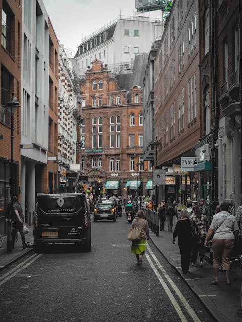 A busy street scene in Soho, London, during daytime, showing pedestrians walking along the pavement in both directions, some carrying shopping bags and backpacks. Several parked and moving vehicles, including a black van with branding for the company 'Man with Van Soho,' are visible on the narrow asphalt road marked with double yellow lines along the edges. The background features a mixture of historic and modern buildings, with ornate brick facades, large windows, and balconies, indicative of typical Soho architecture. On the right side, a storefront with signage and awnings is partially visible, while on the left, the pavement is lined with additional shops and pedestrians. This scene captures the urban environment of a home relocation or furniture transport process, with vehicles likely involved in removals, and pedestrians potentially assisting or observing the moving activities associated with house or apartment moves in Soho.