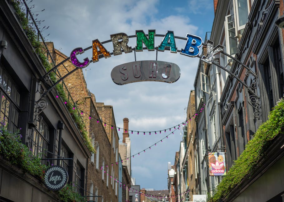 The image shows the entrance to an urban shopping and entertainment area on Soho's Carnaby Street, with the exterior of the Undertbelly venue visible on the left, featuring large glass windows and a brick facade with signage. To the right, there is a narrow pedestrian walkway leading into a passageway framed by a black awning with the name 'artusi,' and several people walking through the street, some carrying shopping bags or backpacks. The area is bustling with activity, and the environment includes modern buildings with large windows, indoor plants visible inside the upper floors, and signs indicating nearby businesses. The scene captures the process of city-based home relocation or furniture transport, with visible movement and the setting for a typical moving or packing scenario outside a commercial property in Soho, where professional movers like Man with Van Soho may operate. Equipment such as bollards and pavement details are also visible, emphasizing the urban context.