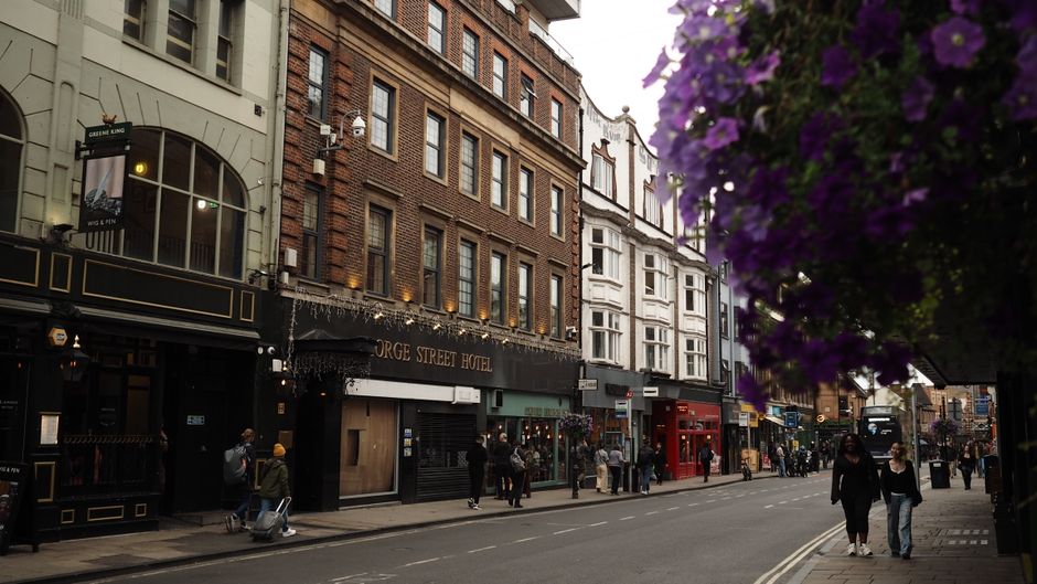 A busy street scene in Soho featuring several multi-storey buildings with brick and white façades, housing various shops and establishments including the George Street Hotel, with signage visible. Th