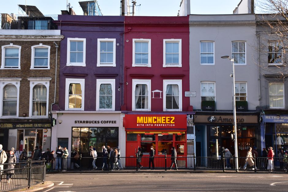 A busy street scene in Soho featuring several multi-storey buildings with brick and white façades, housing various shops and establishments including the George Street Hotel, with signage visible. Th